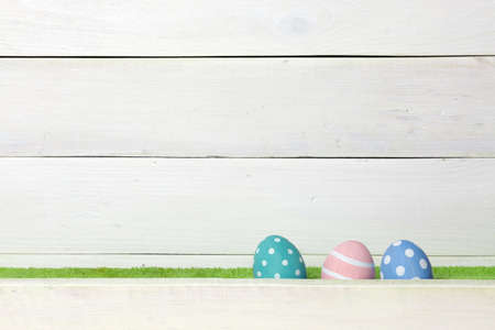 Three colorful handmade easter eggs stand on a green lawn, covered with a barrier, on a white wooden background with space on the leftの写真素材