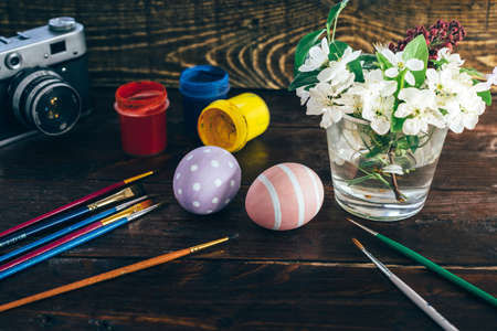 Preparation for Easter. Colorful handmade Easter eggs painted, paints, brushes, retro camera and vase with flowers on a dark wooden backgroundの写真素材