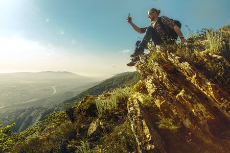 Tourist with  backpack sits on top of the mountain and takes pictures of the landscape on cellphone. Concept of travel, adventure and discoveryの写真素材