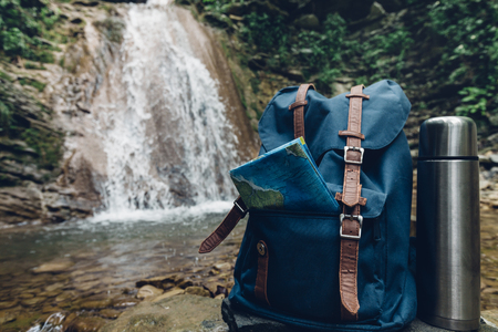Hipster Blue Backpack, Map And Thermos Closeup. View From Front Tourist Traveler Bag On Waterfall Background. Adventure Hiking Conceptの写真素材
