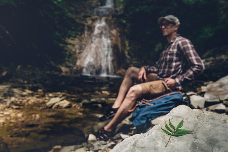 Hiker With Backpack Sits On The River And Enjoys Surrounding View, Selective Focus. Exploring Adventure Hiking Conceptの写真素材