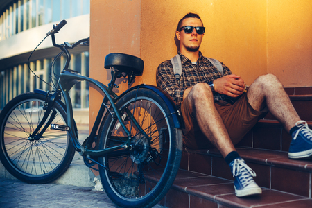 Handsome Guy Cyclist In Sunglasses With Backpack Sitting On The Steps Near The Bicycle Bike And Restingの写真素材