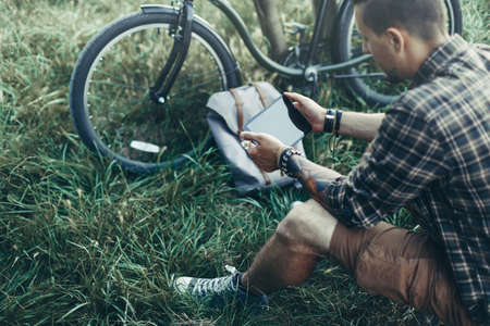 Young Guy Cyclist Sits On Summer Meadow Near Bicycle, Holding And Looking At Tablet Recreation Resting Travel Destination Conceptの写真素材