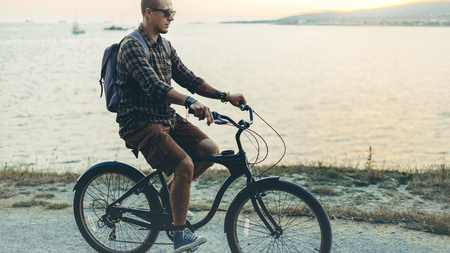 Handsome Young Hipster Man Is Cycling Along The Shore Against The Backdrop Of The Sunset Vacation Holiday Activity Conceptの写真素材