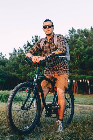 Handsome Young Man Cyclist In Sunglasses With Bicycle In Summer Park During Sunset Vacation Traveling Relaxation Resting Conceptの写真素材