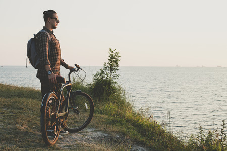 Handsome Young Guy Standing With Bike On Coast And Enjoying View of Nature Sunset Vacation Traveling Relaxation Resting Conceptの写真素材