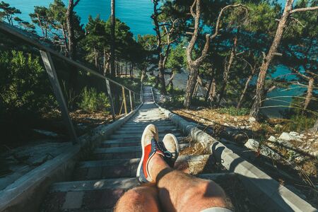 Hiker Man Sitting On Steps Of Stairs Coast Above Sea, View Of Legs. Point Of View Shotの写真素材