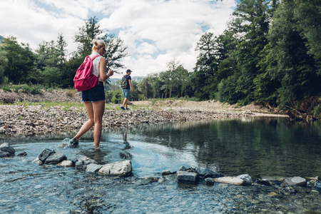 Young Couple Walking Along The Coast Of Mountain River In Summer. Girl Wading Across The Riverの写真素材