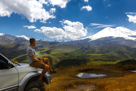 Young Traveler Man Sits On The Sea And Enjoys View Of Mountains In Summer. Elbrus Region, North Caucasus, Russiaの写真素材