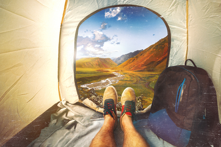 Hiker Man Sitting In A Tourist Tent by The Mountain View With Scratches. Holiday Vacation Concept. View Of Legs. Point Of View Shotの写真素材