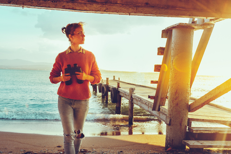 Hiker Girl With Binoculars In Hand Standing By The Sea Near Pier And Enjoying View Of Nature, Front Viewの写真素材