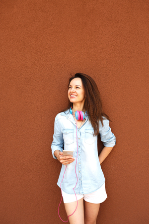 Happy fashionable girl with headphones and a telephone near the wall, smiling and enjoying. Concept of urban style, youth and trends, feminism, emancipation, women's rightsの写真素材