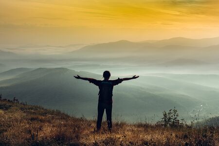 Young girl traveler stands on a cliff in the summer mountains at sunrise and enjoys a view of natureの写真素材