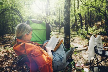 Finding solitude in wilderness concept. Young woman resting in forest, sitting in camp chair and reading bookの写真素材