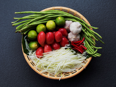 Mix ingredients of Thai green papaya salad (Som tum) composed of shredded papaya, tomato, lime, garlic, chili, and long bean in brown basket on dark backgroundの写真素材