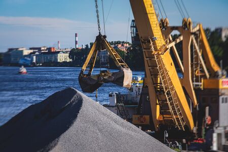 Bulk-handling crane unloading sand, road metal and gravel from cargo vessel ship, heavy vehicle and portal crane loader working with bulk materials in dock terminal の写真素材
