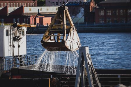 Bulk-handling crane unloading sand, road metal and gravel from cargo vessel ship, heavy vehicle and portal crane loader working with bulk materials in dock terminal の写真素材
