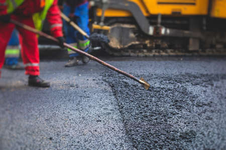 Process of asphalting, blacktopping and paving, group of workers with shovels finishing the asphalt layer, with asphalt paver machine and steam roller compactor vehicle in a backgroundの写真素材