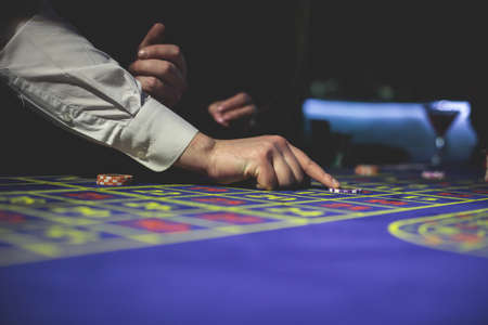 A close-up vibrant image of multicolored casino table with roulette in motion, with casino chips. the hand of croupier, money and group of gambling rich wealthy people playing in the backgroundの写真素材