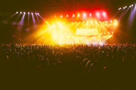 A crowded concert hall with scene stage orange and yellow lights, rock show performance, with people silhouette, colorful confetti explosion fired on dance floor air during concert festivalの写真素材
