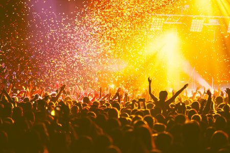 A crowded concert hall with scene stage orange and yellow lights, rock show performance, with people silhouette, colorful confetti explosion fired on dance floor air during concert festivalの写真素材