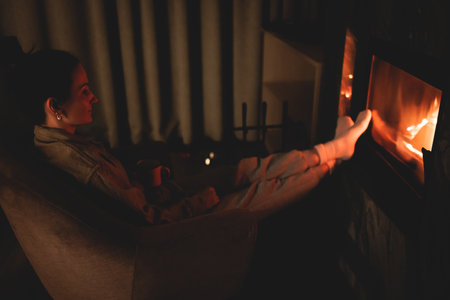Cozy night in the cabin by the fireplace, fireplace burns in the scandinavian cottage chalet house, burning fire with charcoal and firewood, young woman with drink reads a bookの写真素材