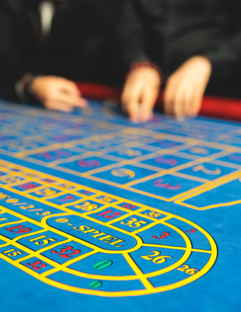 Vibrant casino table with roulette in motion, with casino chips, tokens, the hand of croupier, dollar bill money and a group of gambling rich wealthy people playing bet in the backgroundの写真素材