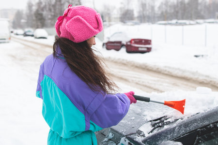 Process of cleaning a car from snow in the morning, girl removing snow from windscreen with a window scraper brush after snowstorm, auto covered in a snow on parking lot, winter timeの写真素材
