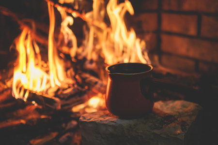 Warming up the fire, cozy winter night in the scandinavian cottage house cabin by the fireplace, burning fire with charcoal and firewood, heating up the drink beverage in a coffee potの写真素材