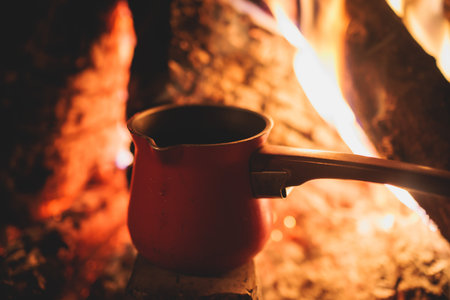Warm by the fire, cozy winter night in the scandinavian cottage house cabin by the fireplace, burning fire with charcoal and firewood, heating up the drink beverage, tea or soup in coffee potの写真素材