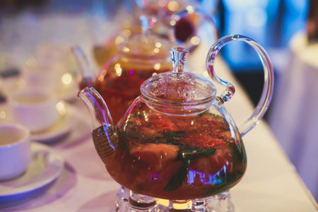 Beautiful warm picture of transparent teapot kettle with tasty green black tea with apple, lemon and ginger on a table with candles and with dessert in the backgroundの写真素材