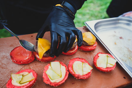 Process of cooking different multi colored burgers on open-air festival, view of chef hands in black gloves with variety of fillings and ingredients on wooden desk and stoveの写真素材
