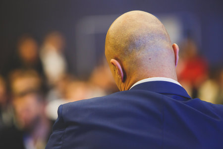A back view of a male Speaker in a blue suit giving a talk on a Stage at a corporate Business...