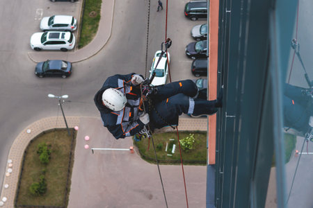 Professional climber rope access worker painting, repairing and cleaning windows on facade of residential skyscraper high rise building exterior wall, industrial mountaineers working at heightsの写真素材
