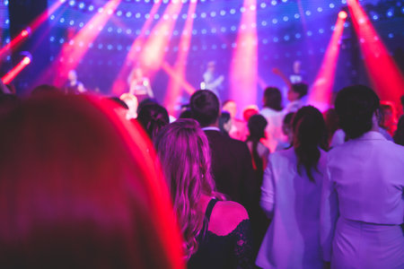 A crowded concert hall arena with scene stage lights with musicians band on a stage at the venue, rock show performance, with concert-goers attendees, audience on dance floor during concert festivalの写真素材