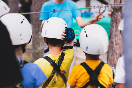 View of high ropes course, kids of climbing in amusement activity rope park, passing obstacles and zip line on heights, children teenagers in equipment gear between the trees on heights, summer dayの写真素材