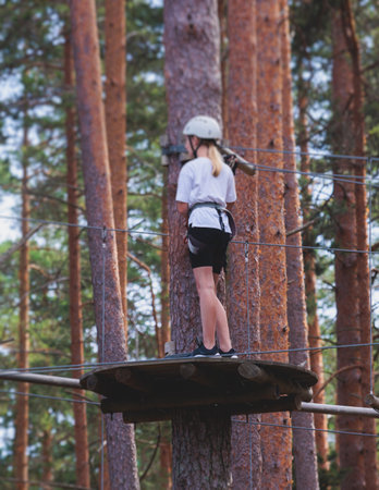View of high ropes course, kids of climbing in amusement activity rope park, passing obstacles and zip line on heights, children teenagers in equipment gear between the trees on heights, summer dayの写真素材