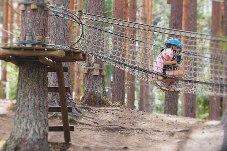 View of high ropes course, kids of climbing in amusement activity rope park, passing obstacles and zip line on heights, children teenagers in equipment gear between the trees on heights, summer dayの写真素材
