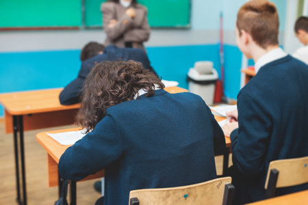 Kids in school writing and taking notes, teens pupils behind desks during the lesson listening to teacher lecture, classroom with students and classmates, group of teenagers during test and examの写真素材
