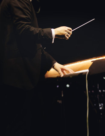 Male conductor behind the podium directing orchestra on stage with pupitre, musicians and choir, the...
