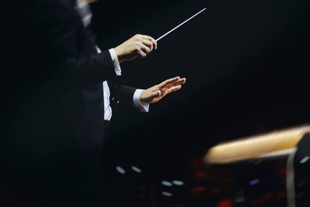 Male conductor behind the podium directing orchestra on stage with pupitre, musicians and choir, the...
