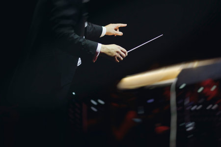 Male conductor behind the podium directing orchestra on stage with pupitre, musicians and choir, the...