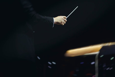 Male conductor behind the podium directing orchestra on stage with pupitre, musicians and choir, the...