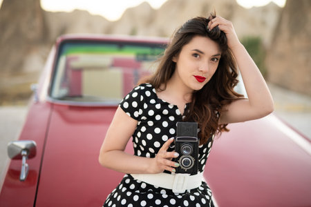 beautiful girl in retro style posing near vintage red cabriolet car with old film camera in handsの写真素材