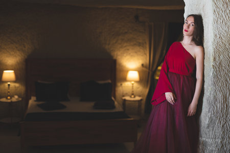 a girl in a red evening dress poses in the interior of a hotel bedroomの写真素材