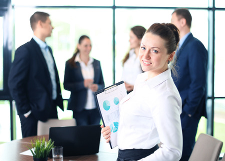 Business woman standing in foreground with a tablet in her hands, her co-workers discussing business matters in the backgroundの写真素材