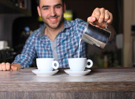 Selective focus on two white cups of coffee and metal jar with milk. Handsome young smiling baristaの写真素材