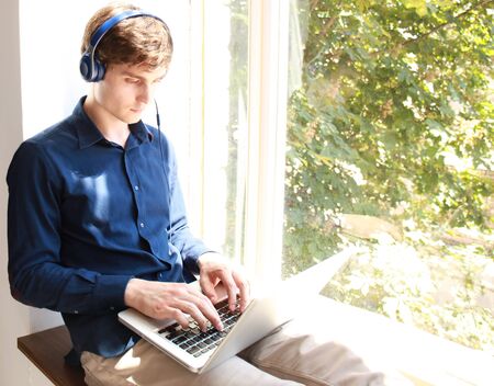 Smiling relaxed young man using laptop with headphones sitting on window sill.