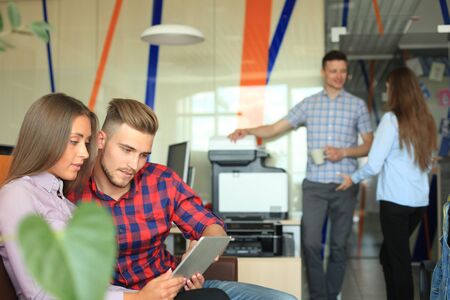 Two confident young people looking at touchpad while their colleagues working in the background.