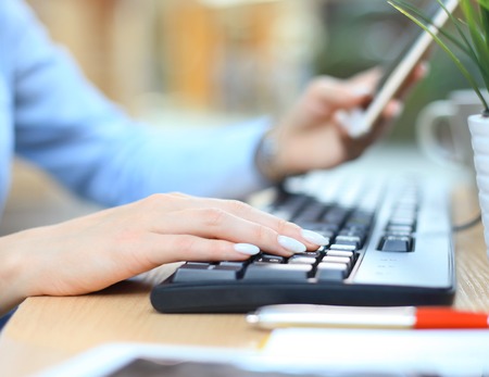 Closeup female hands typing on keyboard using smart phone at the desk in the office.